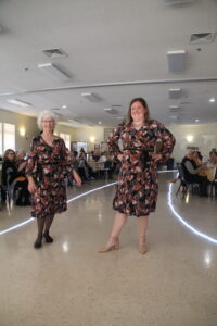 a couple of women walking in a room with people sitting in chairs